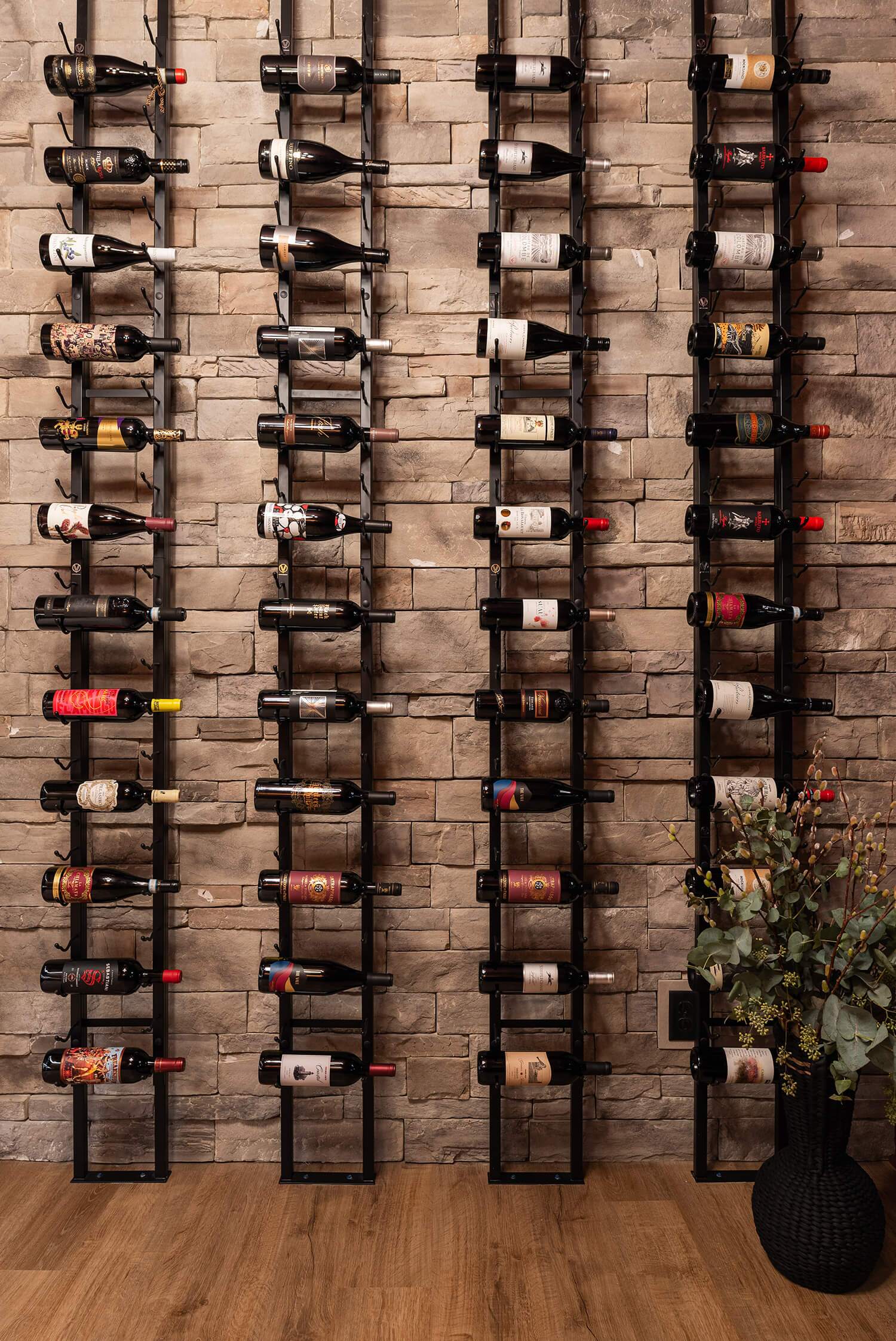 Vertical black racks holding wine bottles on a stone wall, with wood flooring and a black vase of greenery at the side