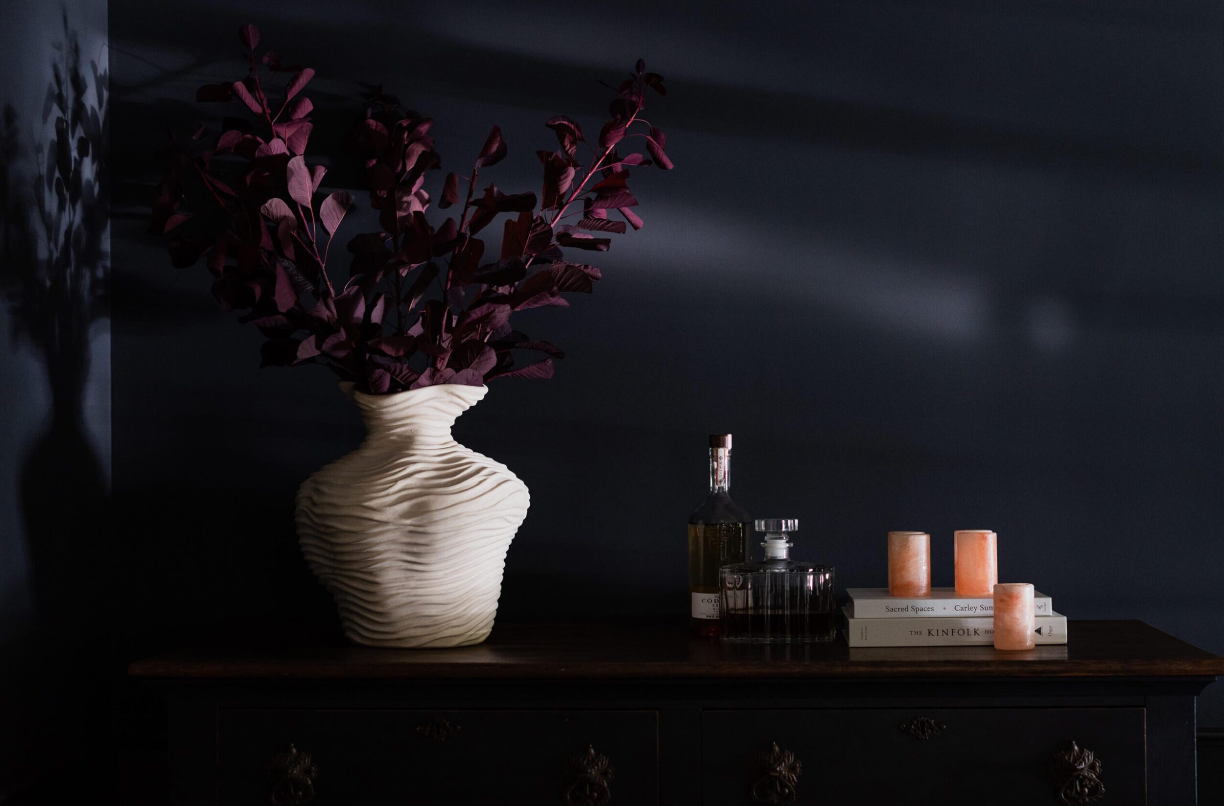 White textured vase with dark foliage, glass decanters, books, and candles on a wooden sideboard.
