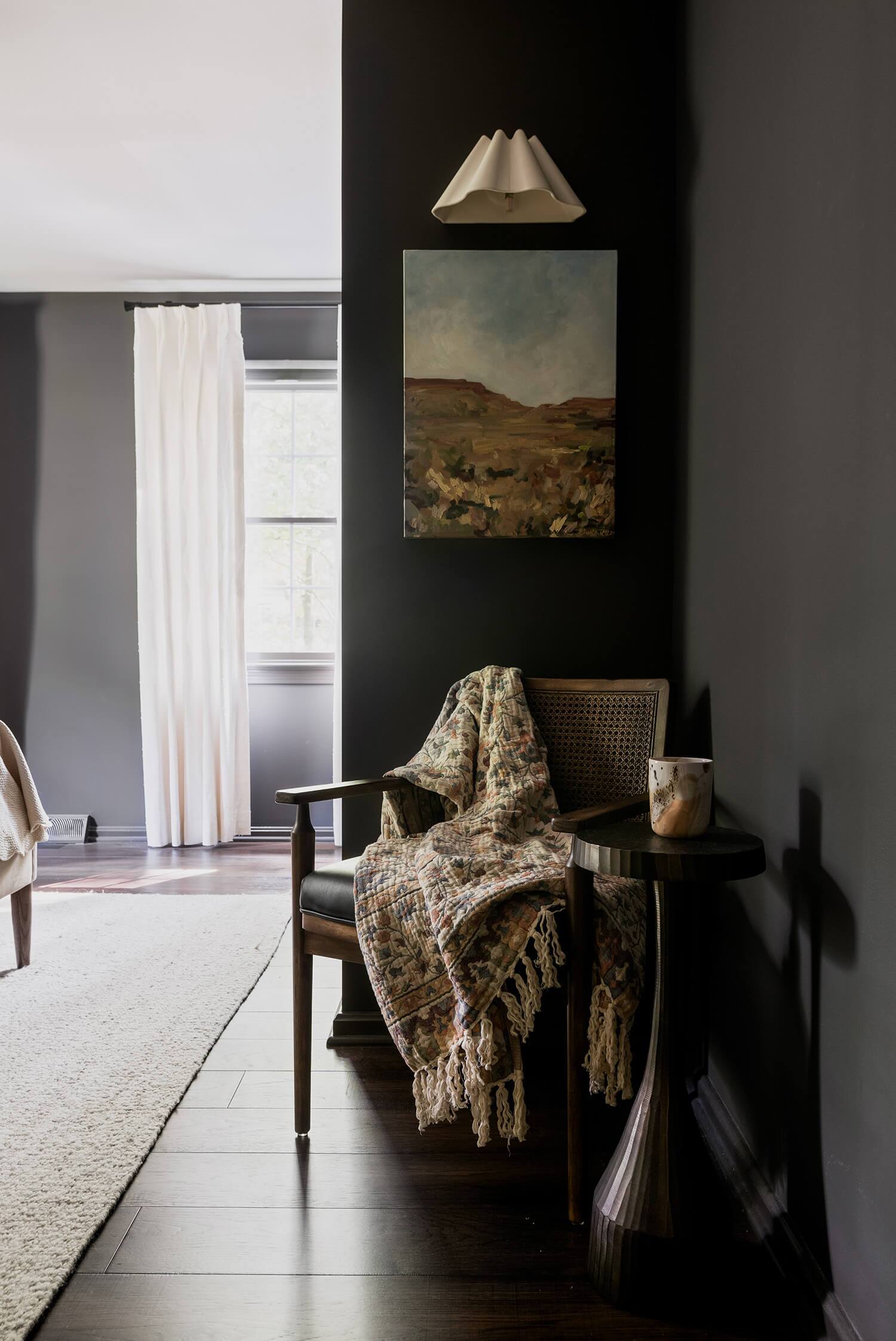 Dark corner with a wooden chair, patterned throw, small side table, and wall art under a light.