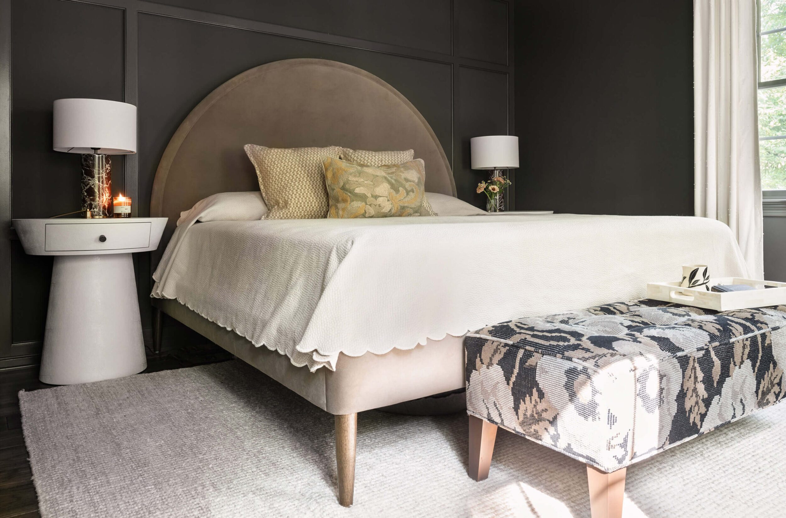 Bedroom with arched headboard, white bedding, side lamps, and patterned bench at foot of bed.