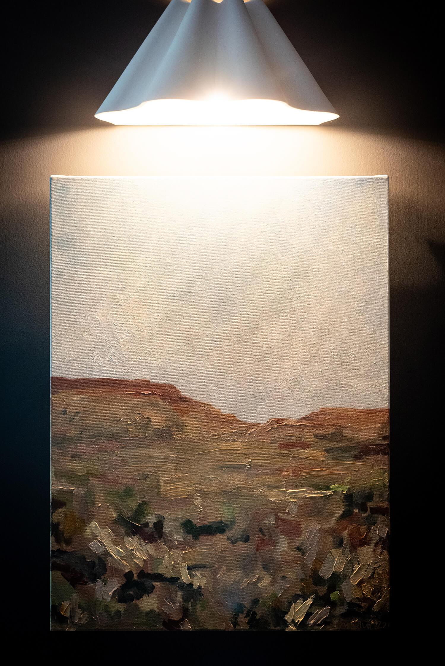 Framed landscape painting with earthy tones, illuminated by a white wall sconce above.