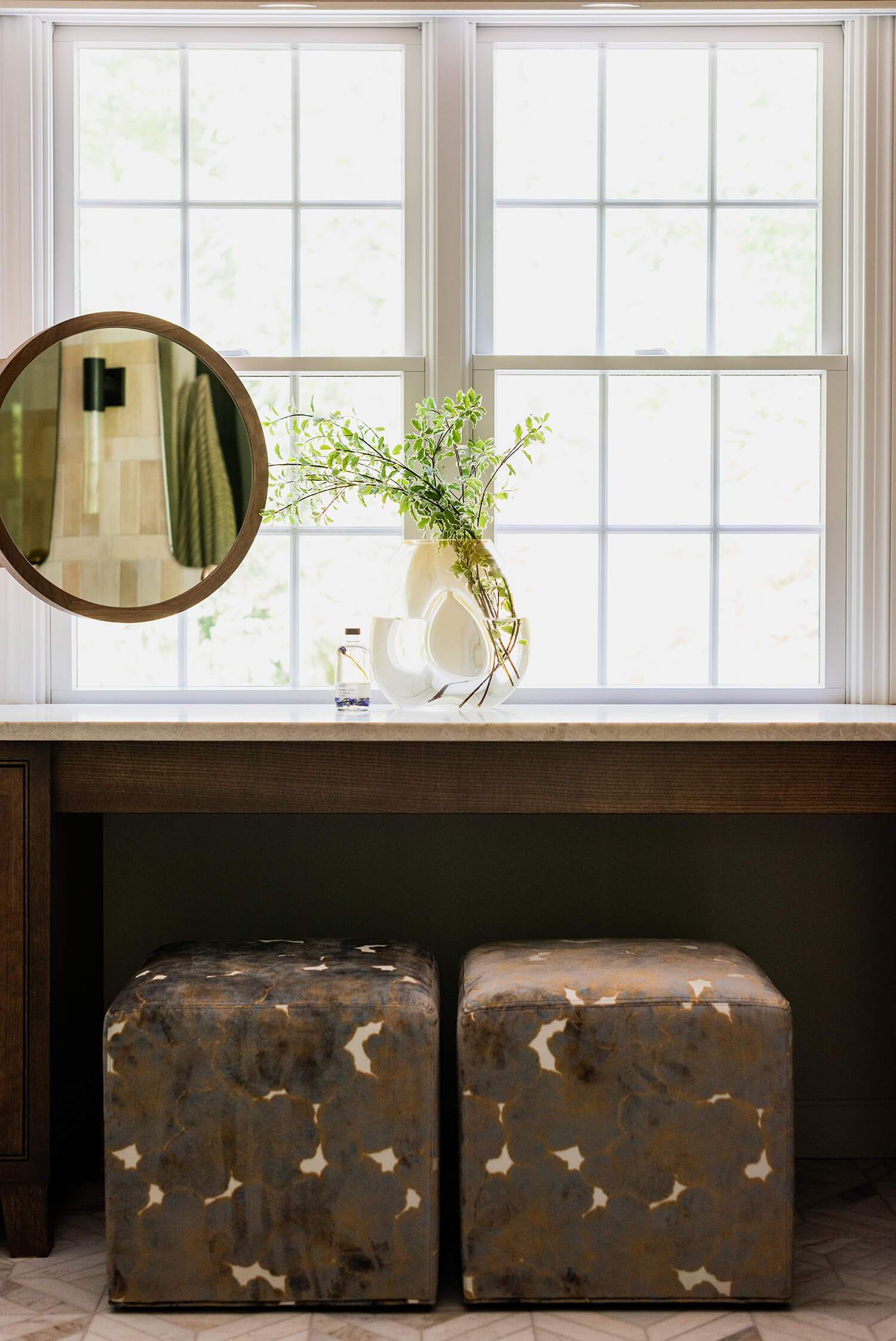 Vanity nook with patterned cube ottomans beneath wood counter, round mirror and vase with greenery before tall sunlit windows