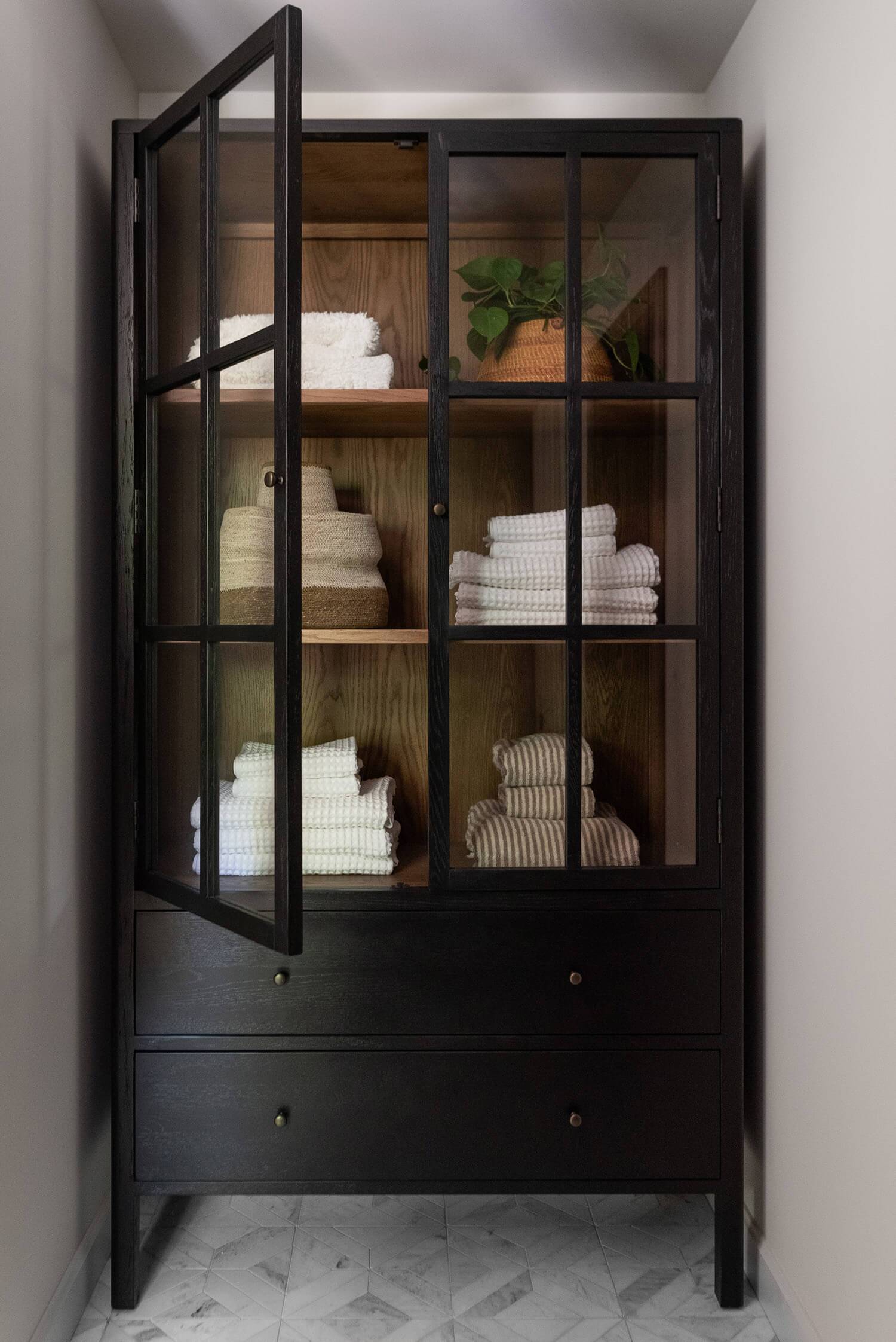 Black glass cabinet displaying folded towels and woven baskets, styled with wood interior and greenery decor