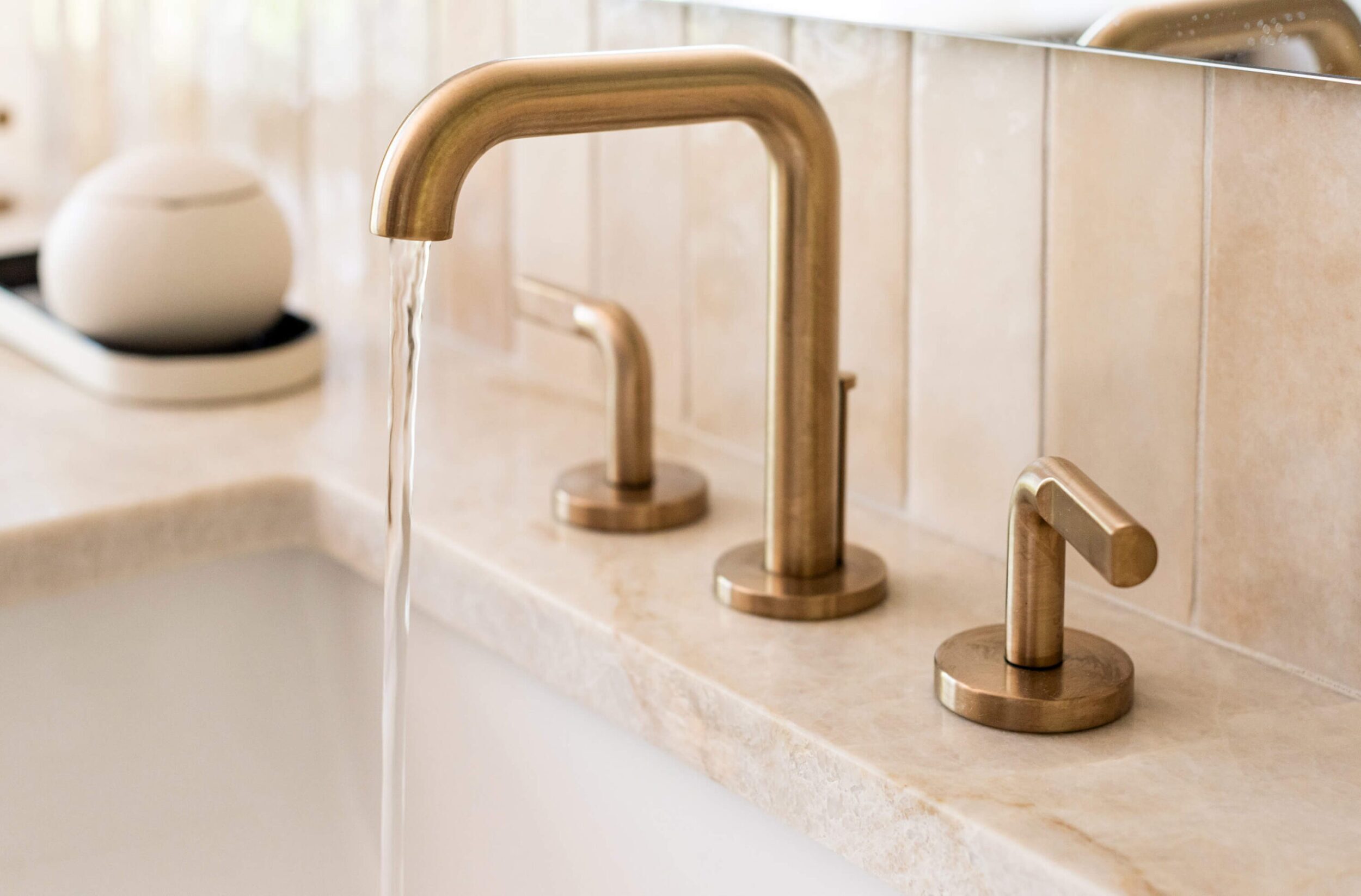 Close-up of modern bathroom sink with brushed gold faucet and running water against a tiled stone backsplash