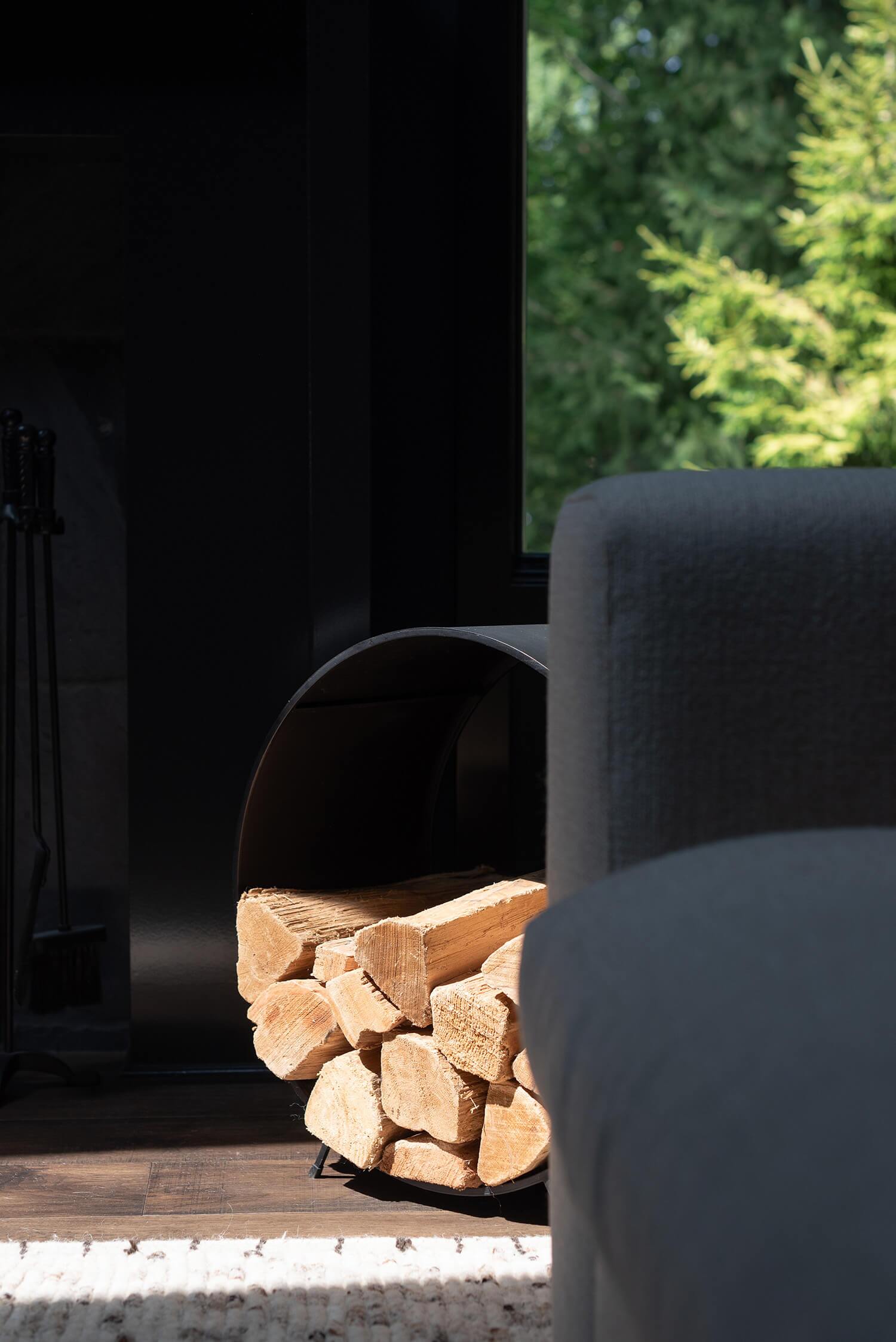 Firewood neatly stacked in black curved holder beside fireplace with view of green trees outside.