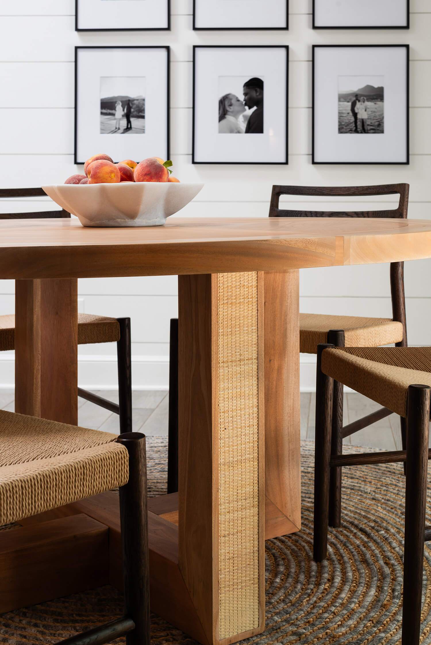 Close-up of round wooden dining table with woven chairs and marble bowl filled with peaches in front of photo gallery wall.