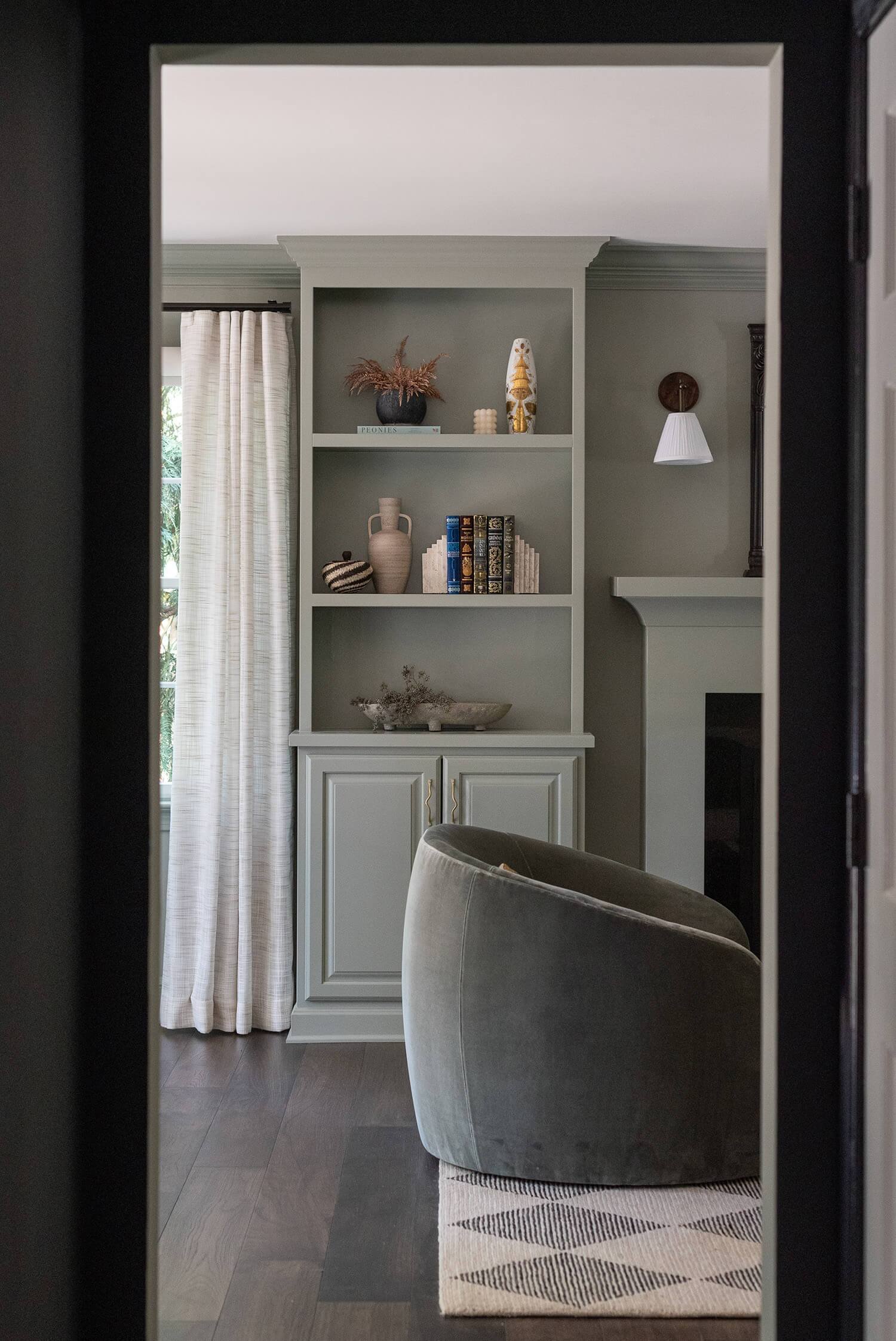 View into a cozy living room with green velvet swivel chair, built-in shelves styled with vases, books, and decor accents.
