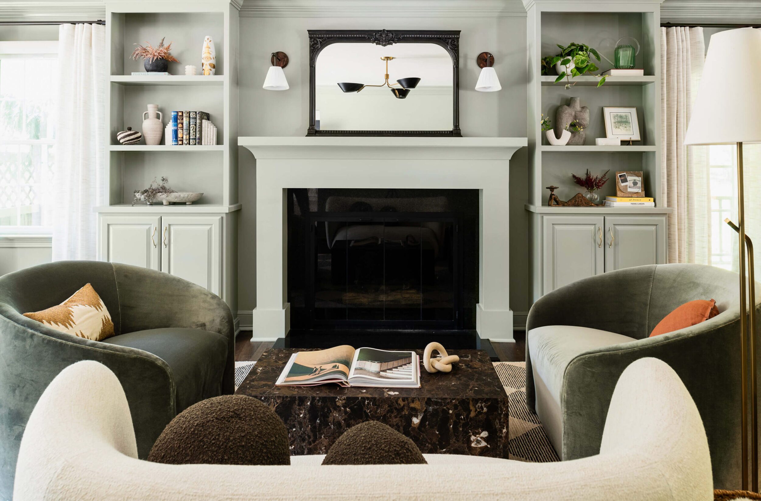 Bright living room with sage built-in shelving, large black fireplace, round velvet chairs, and decorative modern accents.