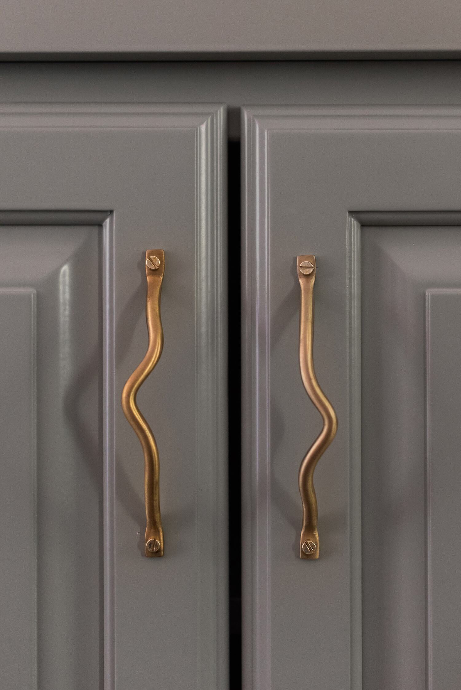 Minimalist cabinet with sage green finish, adorned with unique wavy brass handles in a modern interior detail shot.